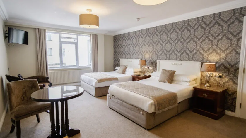 Elegant hotel room with two cosy double beds, ornate wallpaper, soft lighting, seating area, and a flat-screen TV.