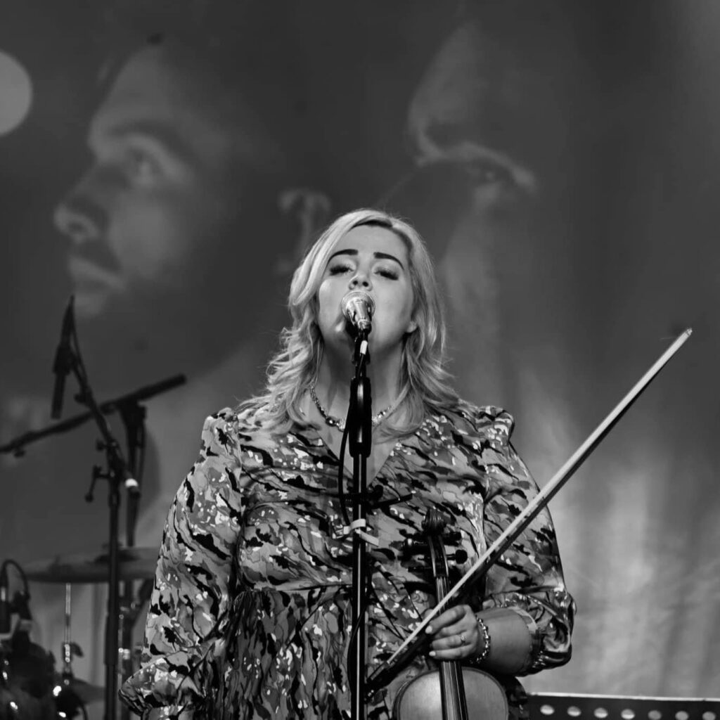 A woman sings into a microphone while holding a violin on stage; two faces are projected in the background.