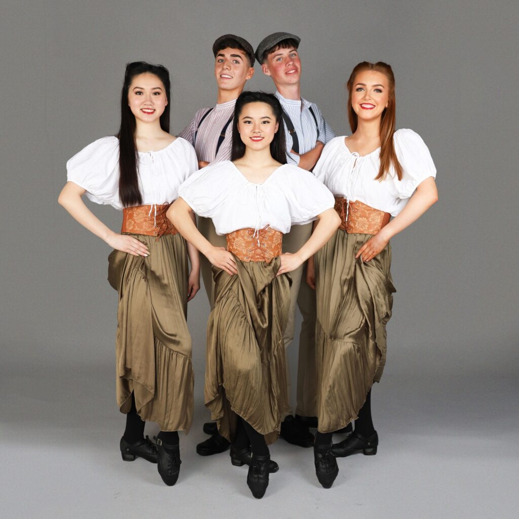 Five young adults in matching costumes, with three women in skirts and white blouses standing in front, and two men in suspenders and caps standing behind, posing against a gray backdrop.