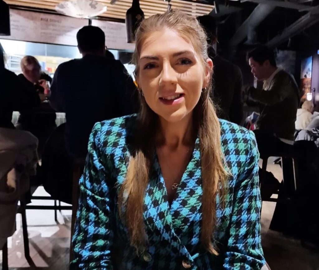 A woman in a blue and black checkered jacket sits at a restaurant table with a plate of noodles and vegetables in front of her.