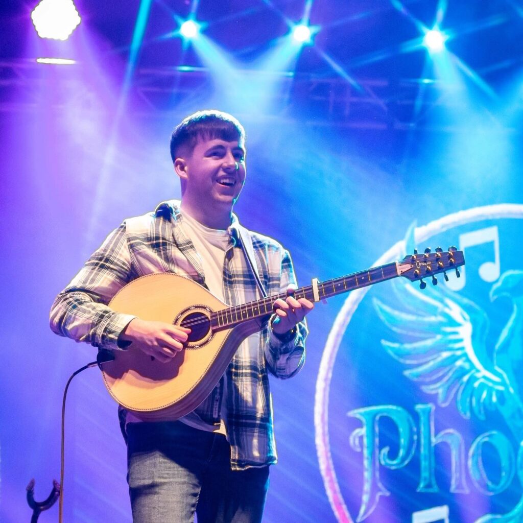 A young man in a plaid shirt plays a mandolin on stage under blue and purple lights, with a circular logo and the word 